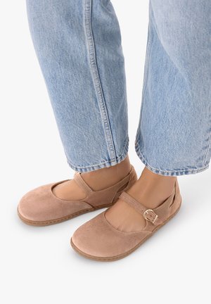 Stopy w jasnoniebieskich dżinsach noszące jasnobrązowe zamszowe buty Mary Jane z paskiem na klamrę na białym tle.