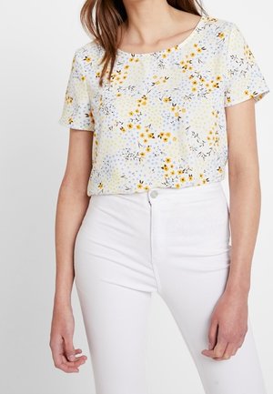 Woman wearing a short-sleeve white top with yellow and blue floral print tucked into high-waisted white pants.