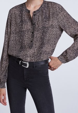 Vrouw draagt een bruin-zwart gevlekt bloesje met lange mouwen, ingestopt in een zwarte high waist jeans met een zwarte riem en zilveren gesp.