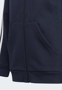 La sudadera con cremallera en color navy presenta una textura suave, dos bolsillos frontales y rayas blancas a lo largo de las mangas. La cremallera tiene herrajes en negro.