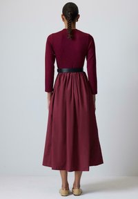 Largo vestido burdeos con un corsé ajustado, mangas largas y una falda acampanada. Cinturón negro en la cintura. Tejido suave con un acabado mate.