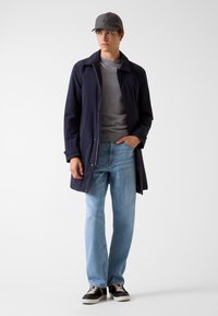 Jonge man met eengrijze pet, marineblauwe jas, grijze trui, lichtblauwe spijkerbroek en zwarte sneakers, die staat met één hand in zijn zak.