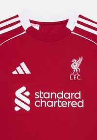 Crvena nogometna majica od prozračnog materijala, s bijelim detaljima, tri bijele pruge na ramenima i logotipima L.F.C.-a i Standard Chartered-a.