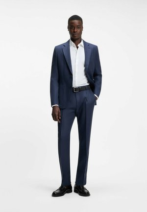 Uomo che indossa un abito blu scuro su misura con camicia bianca, cintura nera e scarpe eleganti nere, in piedi davanti a uno sfondo bianco uniforme.
