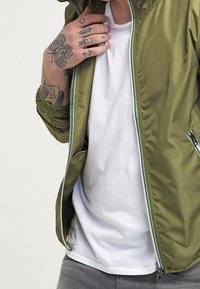 Olivgrön, lätt bomberjacka med dragkedja och fickor. Har en slät, glänsande textur och kontrasterande dragkedjeaccenter.