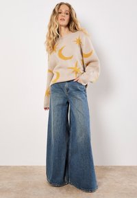 Beige trui met gele maan- en sterpatronen, gecombineerd met wijde blauwe jeans. De trui heeft een ronde hals en ribgebreide manchetten.