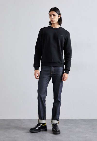 Sort sweatshirt med rund hals, textured stof, parret med mørke denim jeans og sorte snørebåndssko med en tyk sål og gule detaljer.