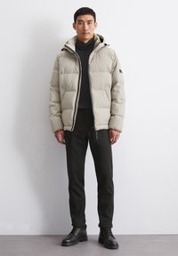 Giacca puffer color beige chiaro con cappuccio, caratterizzata da una chiusura con zip e grandi sezioni trapuntate. Indossata sopra un maglione nero a collo alto e pantaloni scuri attillati.
