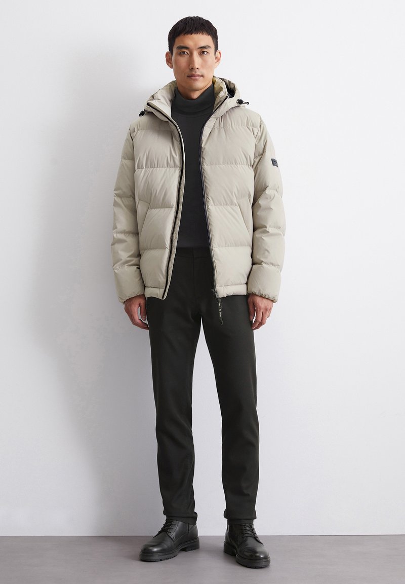 Helle beige Daunenjacke mit Kapuze, mit einem Reißverschluss vorne und großen gepolsterten Bereichen. Getragen über einem dunklen Rollkragenpullover und schlanken dunklen Hosen.