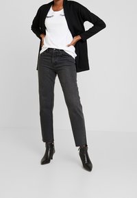 Cardigan negro de manga larga sobre una camiseta blanca con estampado de pestañas, combinado con jeans de pierna recta de tiro alto en gris oscuro y botines negros.