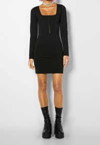 Robe noire à manches longues avec un décolleté carré, une silhouette ajustée, des détails en panneaux et une fermeture éclair à l'avant. Portée avec des bottes plates-formes noires montant jusqu'aux genoux.