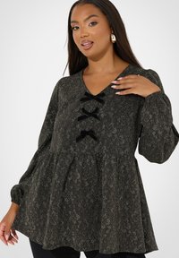 Top nero a fantasia con maniche lunghe, scollatura a V e tre dettagli in velluto nero a forma di fiocco sul davanti. Il tessuto ha una finitura testurizzata e scintillante.