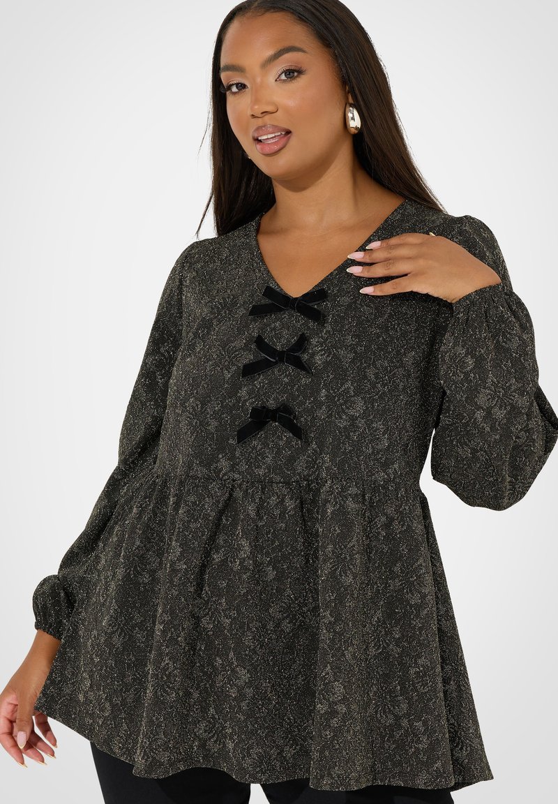 Top nero a fantasia con maniche lunghe, scollatura a V e tre dettagli in velluto nero a forma di fiocco sul davanti. Il tessuto ha una finitura testurizzata e scintillante.