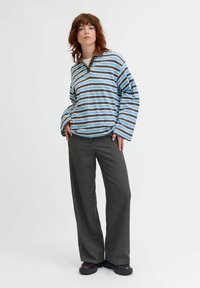 Chemise à rayures à manches longues dans des tons de bleu clair et marron, avec un col à boutons. Associée à un pantalon large gris foncé et des chaussures noires épaisses.