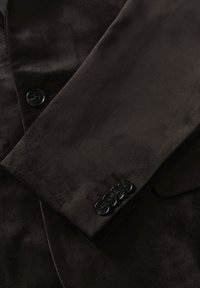 Bruine fluwelen blazer met een zachte textuur, notch revers en vier zwarte knopen aan de mouwen met subtiele stikdetails.