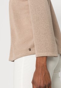 Beige stickad tröjaärm bredvid en mörkhyad hand som vilar vid sidan, tillsammans med vita texturerade byxor.