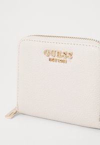 Crème leren portemonnee met gouden "GUESS" logo, ritsluiting en een gestructureerd oppervlak. Bevat een gouden hardware accent op de rits.