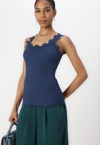 Marineblauw geribde tanktop met golvend kantafwerking, aansluitend ontwerp, zachte textuur, gecombineerd met groene wijde broek, vastgehouden met een blauwe handtas.
