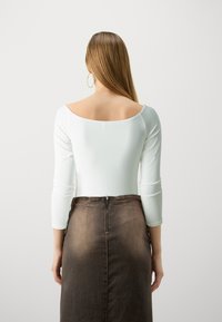 Body alb cu mânecă lungă și spate deschis, purtat cu o fustă din denim maro cu talie înaltă, având un fermoar vizibil la spate.