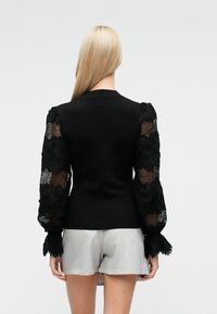 Maglione nero con corpo aderente e maniche in pizzo texturizzato e decorato. Presenta polsini arricciati e collo a costine liscio. Abbinato a pantaloncini grigi.