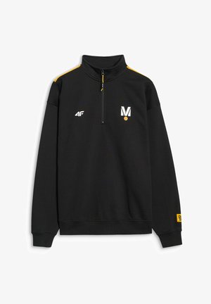 Młody mężczyzna stoi, ubrany w czarny sportowy dres z żółtymi akcentami i czarne trampki, ręce w kieszeniach, neutralne tło.