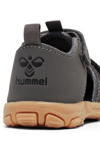 Grå sandal med en glat syntetisk overdel, der har et logo på hælen, justerbar strop og en fleksibel tan gummisål med gripmønstre.