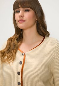 Knapp-up stickad cardigan i krämfärgad med ett mönster, med en orange sammetskant och mörka knappar.