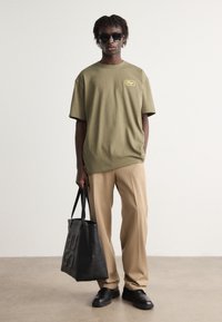 Oliven grøn oversized T-shirt med et lille gult logo, beige bukser, sorte lædersko og en sort tote taske med præget skrift.