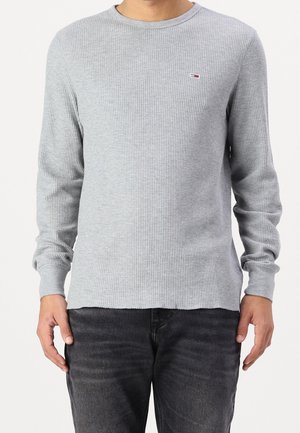 Mand iført lysegrå vaffelstrikket langærmet trøje med lille rød, hvid og blå logo på brystet og mørkegrå jeans.