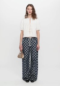 Weiße Bluse mit Puffärmeln und Bindeschnur, kombiniert mit weit geschnittenen karierter Hose in Marineblau und Hellgrau, hält eine kleine gemusterte Tasche.
