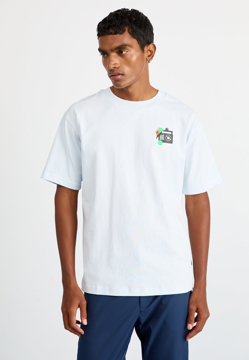 Camiseta de algodón azul claro con mangas cortas, que presenta un gráfico de una cámara en el pecho. Corte relajado, textura suave, diseño sencillo.