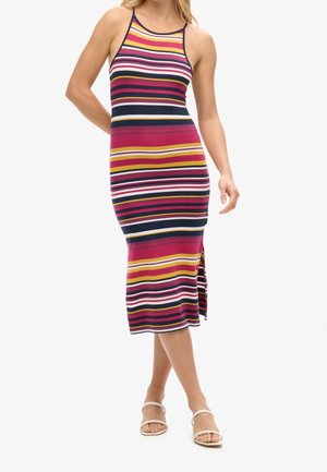 Kobieta stojąca, ubrana w bezrękawnikową, wielokolorową sukienkę midi w paski z bocznym rozcięciem oraz białe sandały z odkrytymi palcami na białym tle.