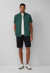 Korte mouwen shirt in het groen met een geometrisch patroon, gecombineerd met zwarte shorts en witte sneakers. Eenvoudige kraag en een relaxte fit.