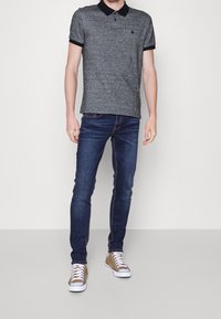 Hombre vestido con una camiseta polo gris con cuello negro, jeans oscuros de corte ajustado y zapatillas color marrón claro con cordones blancos, posando frente a un fondo blanco.