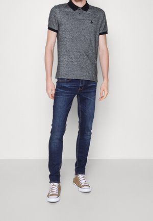 Hombre vestido con una camiseta polo gris con cuello negro, jeans oscuros de corte ajustado y zapatillas color marrón claro con cordones blancos, posando frente a un fondo blanco.