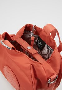 Mała torba z koralowego materiału z zamkiem błyskawicznym w środku, srebrnym zapięciem, w paski podszewką oraz naszywką z logo Kipling z przodu.