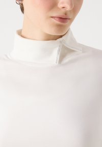 Hvit langermet topp med høy krage som har dekorativ søm og sølvdetaljer. Stoffet har en glatt, lett tekstur.