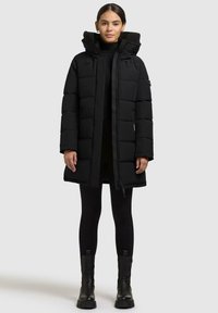 Cappotto imbottito nero con collo alto, texture trapuntata e zip frontale. Indossato su un dolcevita nero e leggings, abbinato a stivali chunky.