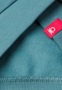 Detailní záběr modrého fleecového materiálu s žebrovanými okraji a červenou štítkem s bílým logem přišitým na švu.