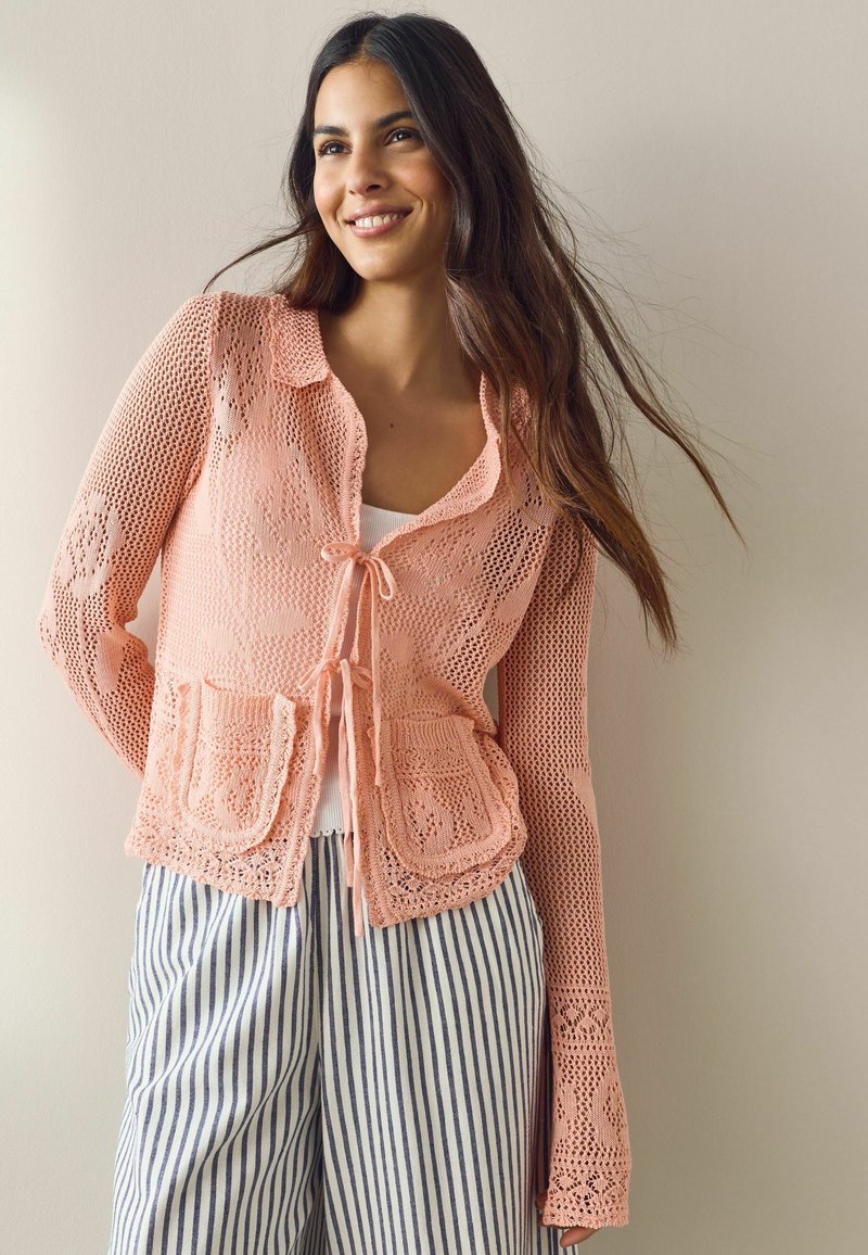 Jeune femme souriante, portant un cardigan en crochet pêche avec des liens à l'avant et des poches, par-dessus un top blanc et un pantalon rayé bleu.