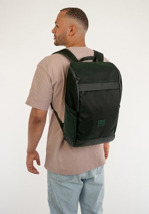 Sac à dos noir de forme rectangulaire, avec des bretelles ajustables, plusieurs poches zippées et un panneau avant texturé.
