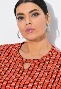 Nainen, jolla on tumma hius, vedetty taakse, käyttää suuria hopeisia korvarenkaita ja puna-mustaa kuvioitua paita, jossa on pieni etuleikkaus.