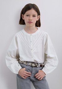 Blusa blanca de algodón con escote redondo y cierre de botones, con costuras decorativas y mangas abullonadas, combinada con jeans grises y un cinturón estampado.