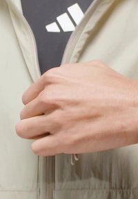Light beige jacket with a zippered front; hand gripping the zipper. Features textured fabric and a black logo underneath.