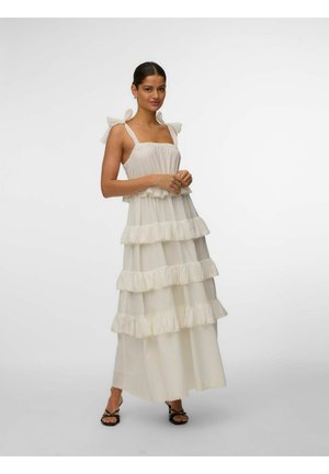 Kvinde iført en ærmeløs, lagdelt maxi-kjole i cremefarve med flæser og båndstropper, stående med hænderne foldet, iført sorte sandaler.