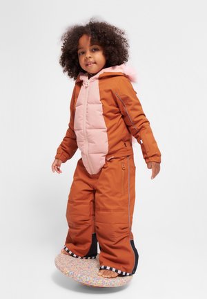 Ett barn i en tvåfärgad brun och rosa vadderad snowboarddräkt med huva och framdragkedja, står på en mönstrad rund surfbräda.
