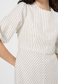 Femme portant une robe blanche à fines rayures verticales noires et manches bouffantes, avec une ceinture où les rayures forment un motif en chevron.