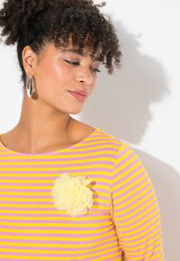 Top a strisce giallo e rosa con scollatura rotonda e un'abbellimento floreale giallo. La modella indossa grandi orecchini a cerchio in argento.