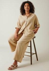 Camicia button-up in lino beige con maniche corte, abbinata a pantaloni larghi coordinati. Piedi in sandali beige. Design semplice e rilassato.