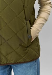 Oliwkowa pikowana kamizelka z teksturowanym wykończeniem, z boczną kieszenią i brązowym wykończeniem dołu, zestawiona z beżową bluzką z długim rękawem o prążkowanej fakturze.